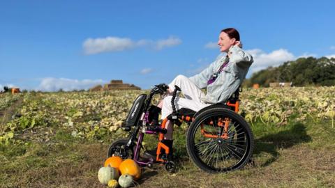 Kate smiling in an orange wheelchair in a field of pumpkins, a bright blue sky. 