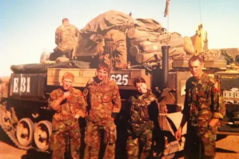 Mel Dyke Mel is in combat gear and posing with three comrades in front of a tank