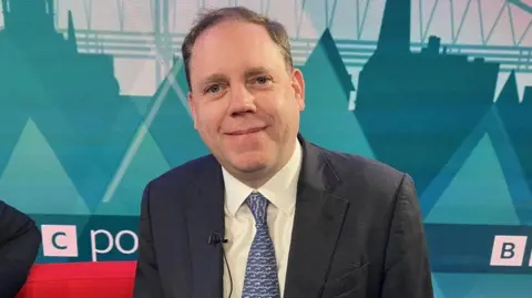 A white man in a grey suit with white shirt and blue patterned tie is seated in a TV studio 