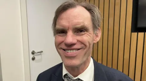 Paul Clarke is smiling in a navy suit with a white shirt and tie. There is a closed door behind him and the all near it has wood panelling.