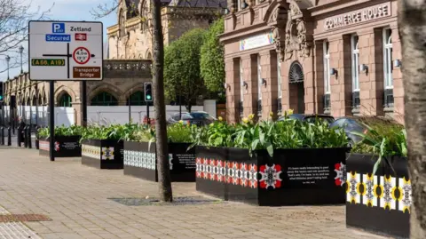 Rachel Deakin A pedestrianised street, with to the right an imposing Victorian sandstone building fronted by raised modern planters with a patterned design on the front and containing flag irises. The ornate frontage of Middlesbrough Station can be seen to the left, with fencing and white barriers around it.
