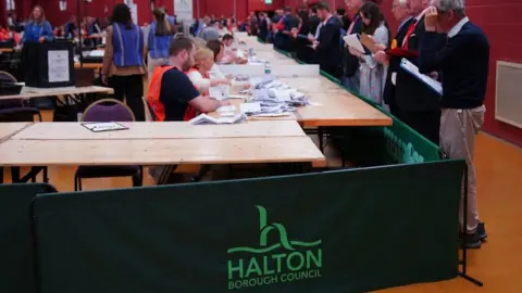 PA Media Rows of tables have been set up with a number of people counting voting slips. There are number of people standing in front of the table overseeing the count. 