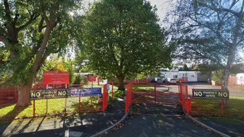 The outside of a primary school. The school has red gates and fences, and three large trees on the grass.