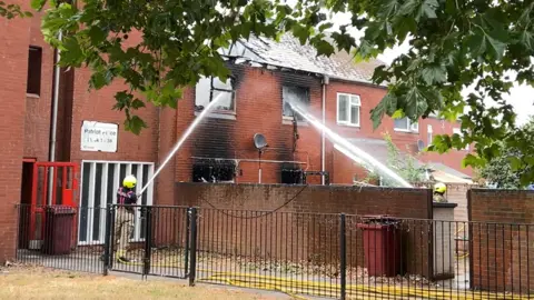 A two-storey house being hosed down with water. Its roof and windows have caved in because of the fire