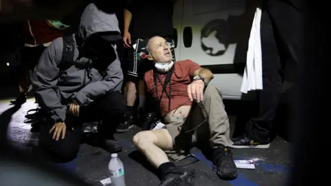 A man wearing a t-shirt, trousers and trainers sits on the pavement, one trouser leg rolled up showing gauze wrapped around his right thigh. He wears a camera around his neck and a respirator mask pulled down. Two people look on to him. 