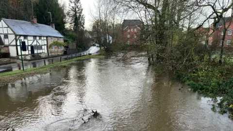 BBC A very swollen river with a road to the left of the image where a large amount of water can still be seen, without houses in the background and a few bare trees