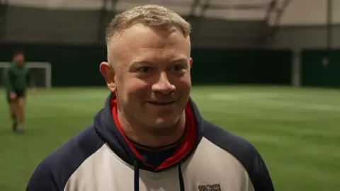 BBC Dan Robinson is a white man, wearing a white hoody, with blue sleeves and good, he has cropped grey hair and is smiling. He's standing on a green all-weather pitch where some of his fellow rugby players can be seen warming-up in the background. 