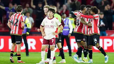 A football player wearing a white Bristol City shirt standing with his arms on his hips in disbelief, while players in red and white shirts celebrate behind him