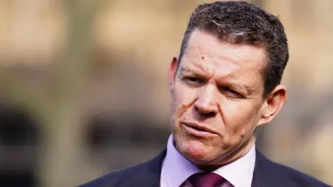 Rhun ap Iorwerth wearing a suit, shirt and tie, speaking in front of a blurred background.
