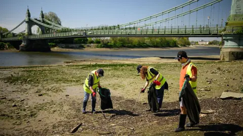 Getty Volunteers can be seen participating in a clean-up of a Thames riverbank. There are three people stood on a beach under a green bridge wearing yellow hi-vis vests putting littler into black bin bags using litter pickers.