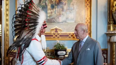 An image showing Grand Chief Joey Pete shaking hands with King Charles III in Buckingham Palace. Behind them is an ornate backdrop featuring gold furniture and a large painting, with light blue walls. Grand Chief Pete (on the left) is wearing a ceremonial indigenous headdress. King Charles is wearing a grey suit. 