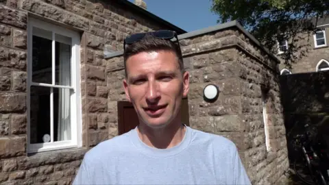 Man in light blue t-shirt with brown hair and wearing sunglasses on his head. He is stood in front of a brown stone building. The sky is blue.