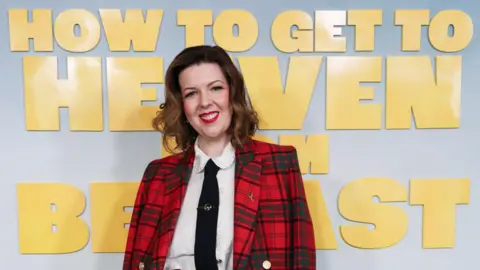 A woman stands in a white shirt and black tie with a red and navy tartan blazer. She has long brown hair and is wearing red lipstick.