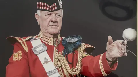 Wally Newman Wally Newman in full Tartan regalia, holding a drum beater, he has a tartan cap and red tunic with gold braiding.
