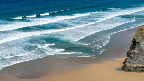 RNLI An aerial photo of a beach on a sunny day. There is a rock surrounded by dry sand which leads to wet sand and waves. The sea is blue and the waves are lined with white but there is a section where the waves do not have the white water. This indicates a rip current.