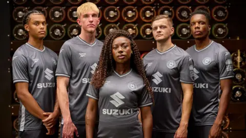 MK Dons A group of people, of different eights and genders are stood in front of the Bombe machine wearing the new MK Dons football kits.