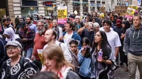 PA Media A group of people, some of whom are holding signs, including some that say 'No Justice No Peace' and 'Black Lives Matter' walk through the centre of Manchester