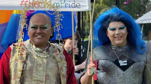 A man in a bright red and gold robe and ornate beaded headpiece takes part in a parade alongside a drag queen with blue hair and a silver sparkly top.