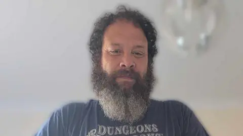 Glen Chisholm A  man with dark curly hair and a large bear looks down at the camera and smiles, he is wearing a blue T-shirt with the word "Dungeons" on it. 