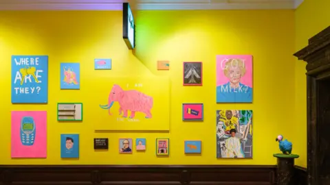 Tom Bird Colourful paintings largely in shades of blue and yellow hang on the yellow walls of an art gallery, with wooden floor. A sculpture of a dodo on a plinth lies to the right corner of the room.