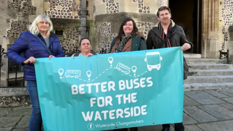 Four people hold up a large blue sign saying "better buses for the waterside" outside a large stone building.