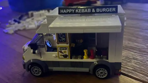 Jo Thompson A kebab van, which has a sign that says Happy Kebab and Burger van above, is stood on a wooden table. It is made out of Lego. 