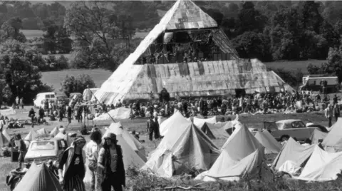 Black and white photo of half built pyramid stage at Glastonbury Festival with rows of tents in front and a car to the left