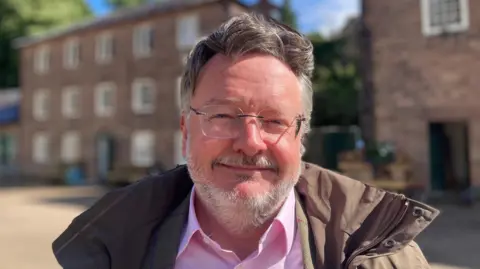 The picture shows Barry Lewis who is the Chair of Derwent Valley Mills World Heritage Steering Group. He is smiling and wearing glasses, with a coat and pink shirt. He is at Cromford Mills in Derbyshire. 