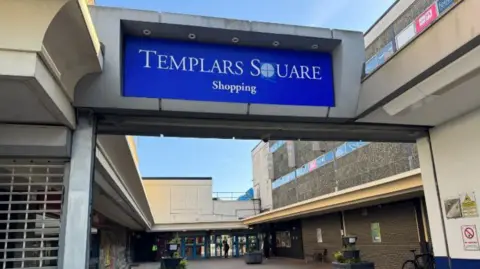 The front entrance to a shopping centre. A large sign above head height reads "Templars Square shopping" in blue, with concrete buildings either side leading to some glass doors. 