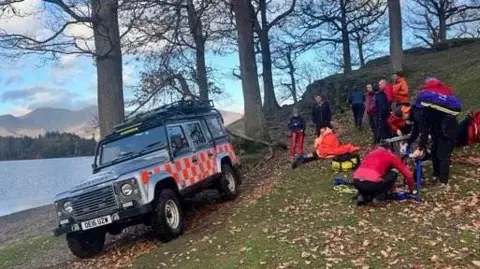 Keswick Mountain Rescue Team Several mountain rescue volunteers - a number of whom are dressed in red tops - attend to a casualty on the path by Otterbield Bay, Derwentwater. There are lots of leaves scattered on the small slope and a mountain rescue vehicle is parked at an angle on the path.