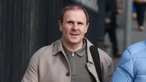 Winston Irvine, a brown-haired man, photographed outside court. He is pictured from the chest up amd wears a olive green zipped up thin jumper under a beige coat. A bag strap is visible on his shoulder. 
