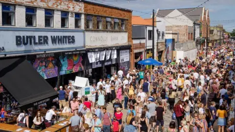 A large crowd on a sunny day in Humber Street, Hull. A series of converted warehouses, now home to restaurants and shops, stand on the far side of the street. To the left is a restaurant with a blue sign reading "Butler Whites" and shutters painted with colourful graffiti.