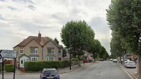 A residential road with houses on either side, parked cars, and a road sign pointing toward a nearby school and community centre.
