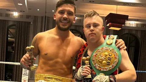 Lee Selby with his arm around Jake Edwards, both wearing boxing gear and in a boxing ring. They are both holding trophies, and Jake has a WBC belt over his shoulder