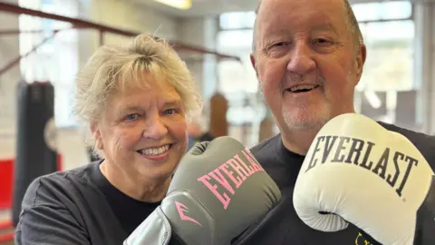 Simon Thake/BBC An elderly couple smile as they hold up their white boxing gloves to the camera.