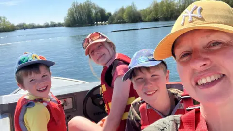 A woman wearing a yellow cap and three children, on a boat. All are wearing life jackets, and the photo is taken as a selfie-style with everyone looking at the camera