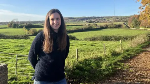 A woman with long, dark coloured hair and wearing a black fleece and blue jeans standing on a wood chipped path in front of open fields.