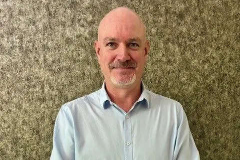 Tom Carter A man smiling with a beard and light blue shirt. There is a patterned light-brown background behind him.