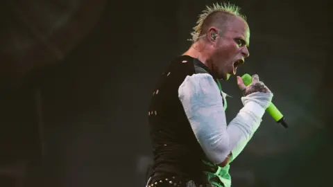 Keith Flint with green spiky hair sings expressively into a microphone on stage. The photo is taken from below, looking up.