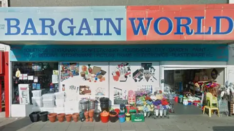 The store has a red and blue sign above it's shop front and goods for sale outside on the ground by the entrance. Goods can also be seen going on into the store.