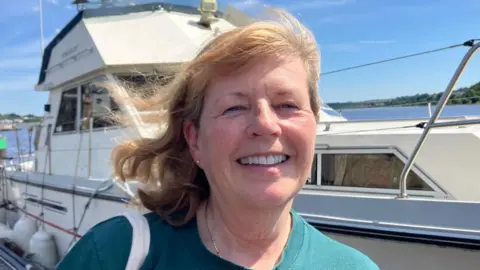 Professor Anne Gilliland stands in front of a boat at the quay in Derry. She is smiling and has long dark blonde hair. Anne is wearing a green t-shirt.