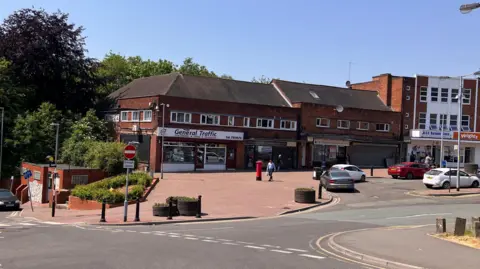 Image shows the end of a shopping street, with some cars parked in a layby in front of shops. The end shop if called 'General Traffic' and has a black and white sign. The other shops next to it are not quite visible as to what they are.