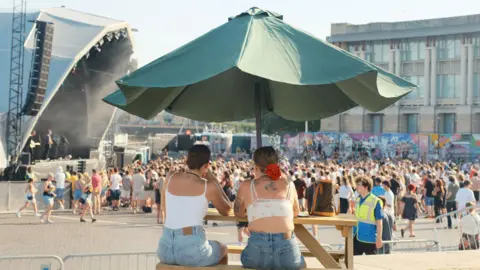 Hayley Thompson Two women are sitting on a bench, under an umbrella with their backs to the camera. They are wearing denim shorts and white tops. They are sat looking over a large crowd of people at a concert. 
