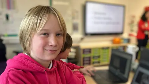 Louis sitting in a classroom, wearing a deep pink hoodie. Other pupils and a whiteboard on the wall can be seen in the background