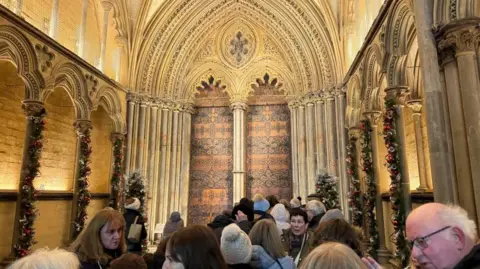 The inside of a cathedral with multiple people inside. Columns inside the cathedral are decorated with green garland and baubles.