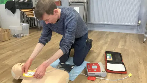 BBC/Katie Radley A man is kneeling on a blue mat on the floor over a mannequin. He is placing a pad on the chest of the mannequin with red medical equipment and a red case on the right hand side. Behind him are stacked chairs and a boxes.