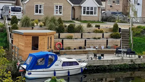 Tony Gosling A 4.5m long (15ft) white cabin cruiser with a blue canopy. There is a grey and orange outboard motor at the back and the vessel is moored to a wooden decked area with a wooden summerhouse. Patio furniture, tables and chairs are on the right and there is a staggered raised bed effect leading up to the back garden of a modern bungalow with other homes around it.