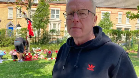Marc Gaier/BBC John Barlow, who has light grey hair and is wearing glasses and a dark blue Adidas branded hoodie, looks into the camera with a solemn expression while in the backdrop a man lays flowers by a tree surrounded with tributes. 