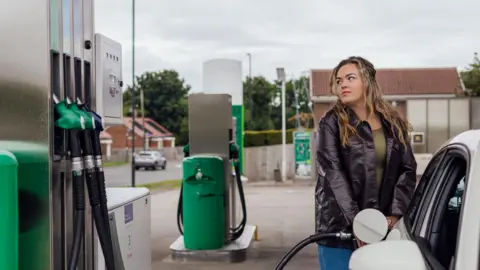 A woman with long brown hair with blonde highlights filling her car up at a petrol station.