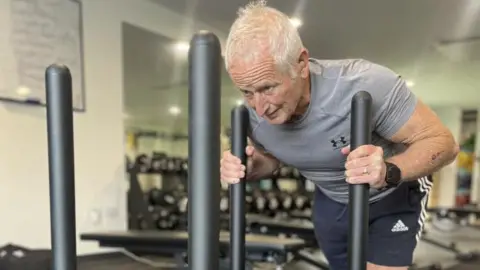 Peter Herbert in a grey tight-fitted shirt and blue shorts. His arms are grasping two vertical bars on an exercise sled which are almost touching his shoulders. His head is down and he is straining in his face and neck. He has white hair. 